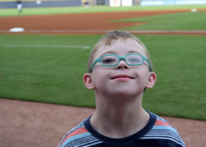 Closeup of a child with Down syndrome wearing glasses with a baseball field in the background.
