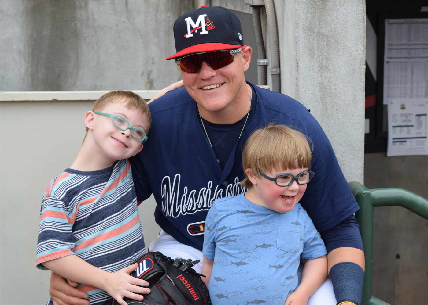 Two children wearing glasses with Down syndrome pose with an older baseball pitcher in front of a dugout.