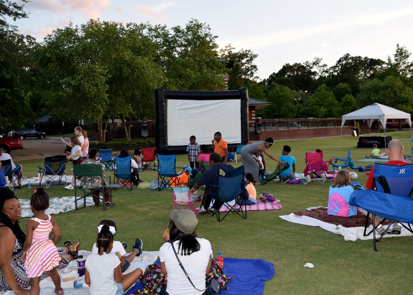 Many families sit with their backs to the camera on the ground and in folding chairs and blankets outdoors and face a large movie screen in a park setting.